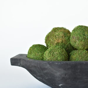 May include: A black wooden bowl filled with green moss balls. The bowl has a unique shape with a handle on one side.
