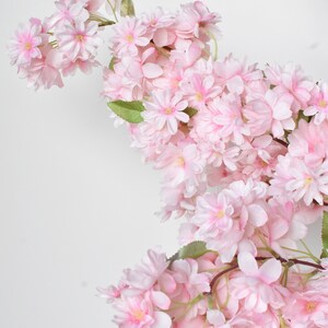 Puede incluir: Una cascada de flores de cerezo artificiales en tonos rosas suaves. Las flores tienen m&uacute;ltiples capas de delicados p&eacute;talos y un centro amarillo, con hojas verdes intercaladas. Las ramas son de color marr&oacute;n natural, sobre un fondo blanco.