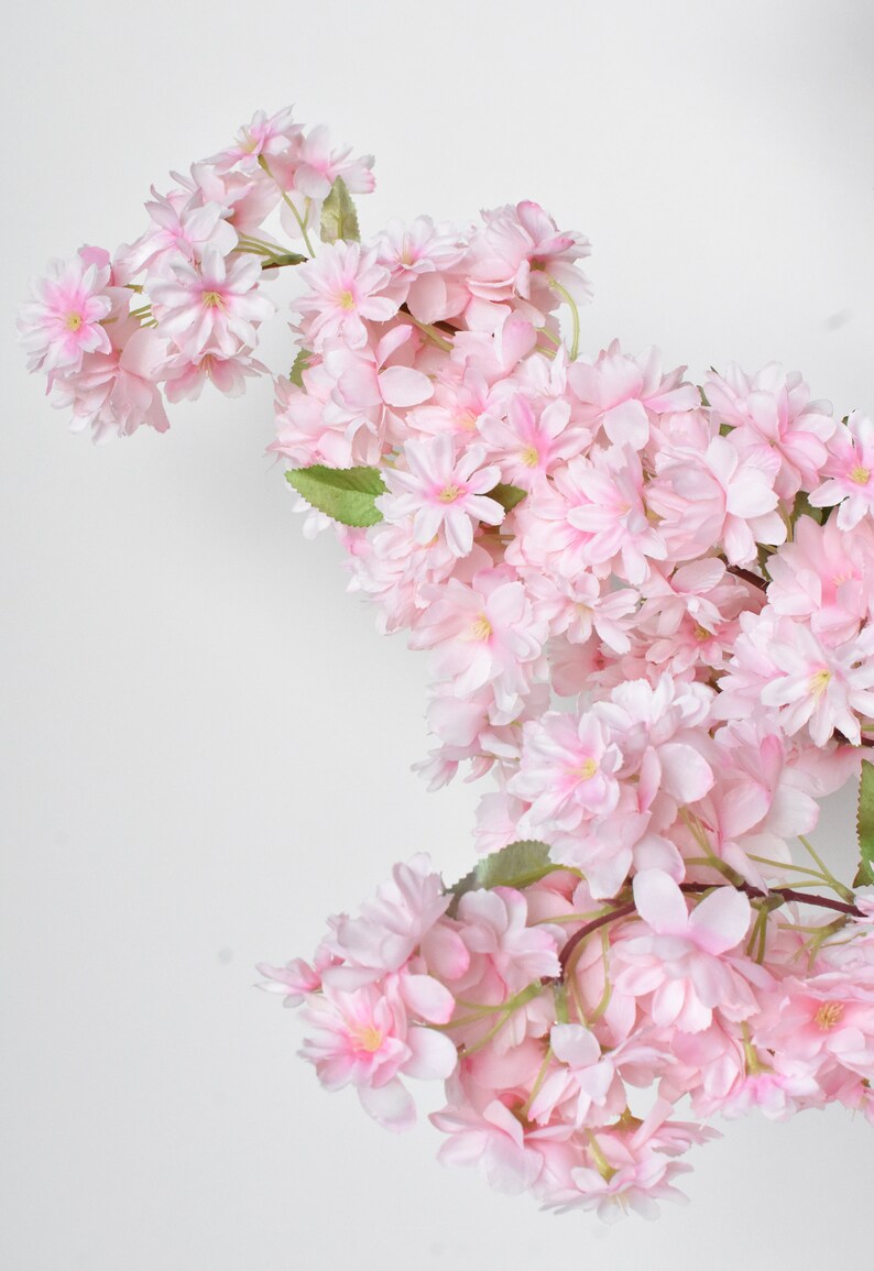 Puede incluir: Una cascada de flores de cerezo artificiales en tonos rosas suaves. Las flores tienen m&uacute;ltiples capas de delicados p&eacute;talos y un centro amarillo, con hojas verdes intercaladas. Las ramas son de color marr&oacute;n natural, sobre un fondo blanco.