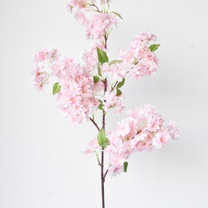 Puede incluir: Un solo tallo de flores de cerezo artificiales. Las flores son de un rosa suave, agrupadas a lo largo del tallo marr&oacute;n con hojas verdes. Las flores est&aacute;n en plena floraci&oacute;n, creando un aspecto delicado y natural. Ideal para la decoraci&oacute;n del hogar.