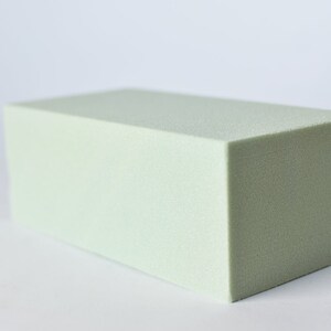 May include: A rectangular block of light green floral foam. The foam has a slightly textured surface and is cut into a precise, geometric shape. The block is positioned on a white background.