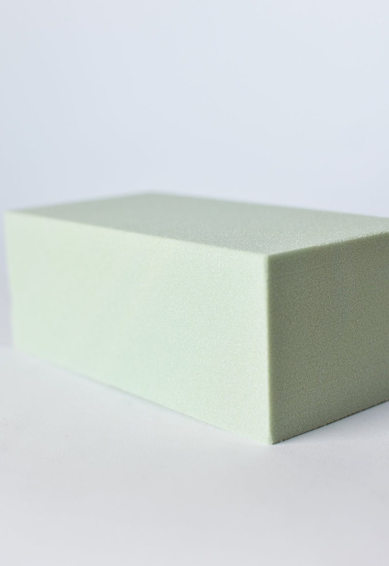 May include: A rectangular block of light green floral foam. The foam has a slightly textured surface and is cut into a precise, geometric shape. The block is positioned on a white background.