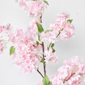 Puede incluir: Una rama de flores de cerezo artificiales con racimos de flores de color rosa p&aacute;lido y hojas verdes. Las flores est&aacute;n en plena floraci&oacute;n, con p&eacute;talos delicados y centros amarillos. El tallo es de color marr&oacute;n oscuro, sobre un fondo blanco.
