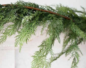 May include: A close-up of a green artificial cedar garland with a brown stem. The garland is draped over a white surface. The garland has many small, detailed, green leaves.