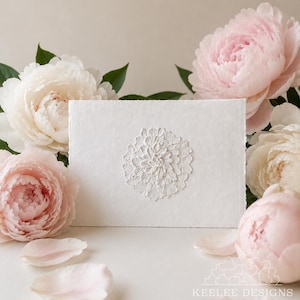May include: A rectangular white card with a deckled edge featuring a central raised white floral relief design. The card is surrounded by several soft pink and white peony blossoms and scattered petals, with the text KEELEE DESIGNS in white.