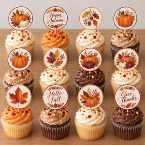 Könnte beinhalten: Ein Set aus zwölf Cupcakes mit runden Papieraufsteckern, die herbstliche Motive wie Kürbisse, Herbstblätter und Eicheln zeigen, sowie Schriftzügen wie Happy Autumn, Hello Fall und Give Thanks, angeordnet in einem Raster auf einer Holzfläche.