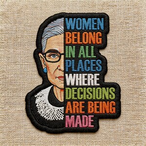 May include: Embroidered patch featuring a portrait of a woman with glasses and the text "WOMEN BELONG IN ALL PLACES WHERE DECISIONS ARE BEING MADE" in various colors. The patch has a black border and is on a tan fabric.