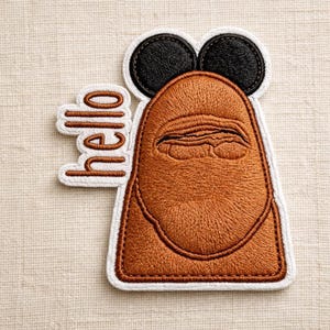 May include: Embroidered patch featuring a brown, stylized face with closed eyes and two black circles above. The word "hello" is embroidered in a cursive font to the left of the face. The patch has a white border.