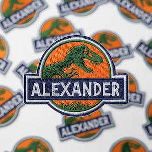 May include: Embroidered patch featuring a green dinosaur silhouette on an orange background, with the name "ALEXANDER" in white letters on a navy blue banner. The patch has a white border and is surrounded by similar patches.