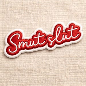 May include: A red and white embroidered patch with the word "Smut Slut" in a cursive font. The text is in red with a white outline and a white border. The patch is on a beige fabric background.
