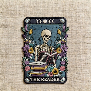 May include: Embroidered patch featuring a skeleton reading a book, surrounded by flowers and a crescent moon design. The patch has the words "THE READER" at the bottom. The color palette includes blue, purple, yellow, and white.