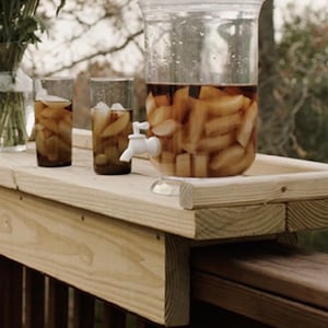 May include: A wooden deck table holds a clear beverage dispenser with a white spigot, two glasses with ice and a vase of flowers. The dispenser is filled with a light brown liquid and fruit slices.