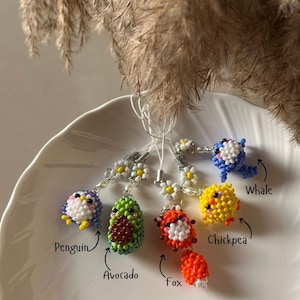 May include: A collection of beaded charms in the shape of a penguin, avocado, fox, chickpea, and whale. Each charm is made with colorful beads and attached to a daisy chain. The penguin is blue, white, and yellow. The avocado is green and brown.
