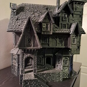 Könnte beinhalten: Ein detailliertes, dunkelgraues Miniatur-Spukhaus mit mehreren Etagen, Giebeldächern und einem markanten Turm. Das Haus verfügt über aufwendige Fensterdesigns, einen Schornstein und eine steinartige Fassade, die eine gruselige, gotische Ästhetik erzeugt.