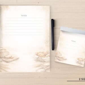 May include: A large notepad and a smaller notepad, both featuring a beige watercolor illustration of bread, wheat, and flour at the bottom. Each notepad has the word NOTES printed at the top above horizontal lines. A black pen sits between them.