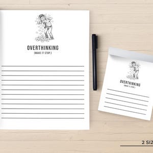 May include: A white notepad and a larger lined paper pad with the text "OVERTHINKING [MAKE IT STOP.]" and an illustration of a person. A black pen rests beside the notepad. The image is on a light brown wooden surface.