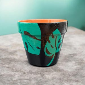 Puede incluir: Una pequeña maceta de terracota pintada a mano. La maceta presenta un diseño de hojas verde turquesa con detalles marrones sobre una base negra. El borde interior está pintado de naranja. Adecuada para plantas pequeñas y añade un toque decorativo.
