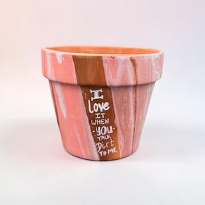 May include: A hand-painted terracotta flower pot with a pink, white, and brown marbled design. The pot features the text "I love it when you talk dirt to me" in white lettering, perfect for planting flowers or herbs.