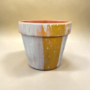 May include: A small, hand-painted flower pot with a white, gold, and peach color scheme. The exterior features vertical streaks of white and gold paint, while the interior rim is peach. This decorative pot is suitable for small plants.
