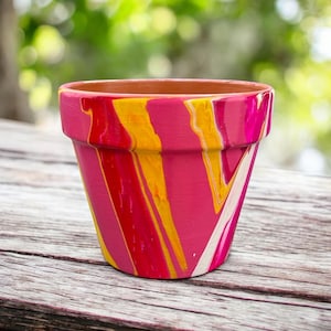 Puede incluir: Una pequeña maceta de terracota con un diseño abstracto vibrante. La maceta está pintada con rayas verticales de color rosa brillante, amarillo y blanco, creando un patrón colorido y llamativo. El borde y el interior son del color terracota natural.