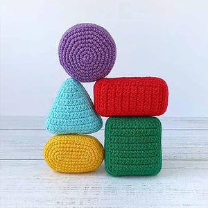 Op de afbeelding: Een stapel kleurrijke, gehaakte geometrische vormen. De stapel bevat een paarse bol, een rood rechthoekig prisma, een groene kubus, een blauwe driehoek en een gele ovaal. De objecten zijn gerangschikt op een wit houten oppervlak.