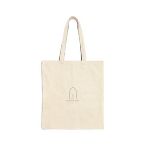 Peut inclure: Sac fourre-tout crème avec de longues anses. Le sac présente un design minimaliste avec une simple illustration d'une fenêtre cintrée et le texte "QUIET IS HOLY" en police fine et foncée. Le sac est fait d'un tissu naturel et robuste.