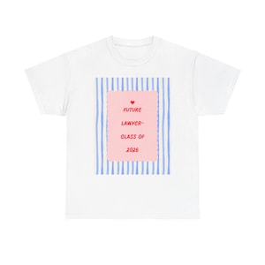Peut inclure: T-shirt blanc avec un rectangle rose portant l'inscription "FUTURE LAWYER - CLASS OF 2026" et un petit cœur rouge. Le rectangle est placé sur un fond de rayures verticales bleues.