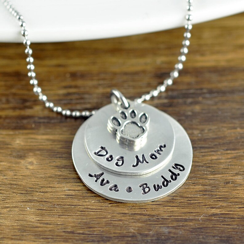 May include: Silver necklace with a layered pendant featuring a paw print charm, the words "Dog Mom" and "Ava & Buddy".
