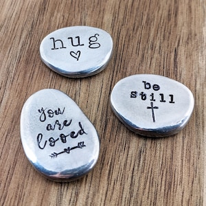 May include: Three small, silver-toned, oval-shaped stones with inspirational messages engraved on them. The stones read "hug" with a heart, "you are loved" with an arrow and heart, and "be still" with a cross.