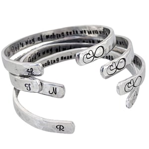 May include: Four silver-toned cuff bracelets. Each bracelet has a hammered texture and an open design. Some bracelets feature an infinity symbol and engraved text. One bracelet has the letter "L", another has "S", "N", and "R".