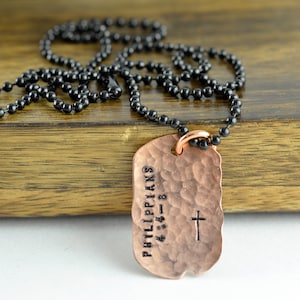 May include: A copper dog tag necklace with a black chain. The dog tag is hammered and has the text "Philippians 4:13-8" and a cross engraved on it.