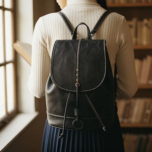 Può includere: Zaino in pelle nera con chiusura a patta e cuciture bianche. Presenta spallacci regolabili e chiusura con coulisse. Il design include una catena di perline decorativa e una piccola maniglia. Indossato da una persona con un maglione bianco.