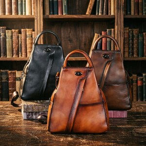 Puede incluir: Tres mochilas de cuero en negro, marrón y beige, cada una con un asa superior y un bolsillo delantero. Las bolsas se exhiben sobre una superficie de madera frente a una estantería llena de libros antiguos. Las bolsas tienen un diseño vintage y clásico.