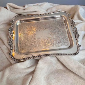 May include: A rectangular silver-toned serving tray featuring an ornate, embossed floral pattern across the center surface. The edges of the tray are decorated with intricate scrollwork and a rope-style border detail along the perimeter.