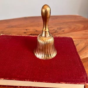 Pode incluir: Uma campainha de m&atilde;o em metal dourado com um cabo liso e alongado e um corpo canelado em forma de sino. O sino apresenta estrias verticais na sua superf&iacute;cie exterior, criando um aspeto texturado. Est&aacute; posicionado na vertical sobre um livro vermelho.