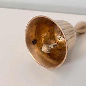Pode incluir: Uma campainha de m&atilde;o de metal em tom dourado com um design exterior canelado. O interior da campainha apresenta um badalo suspenso por um fio e um pequeno autocolante que diz Made in India. A campainha tem uma pega lisa no topo.