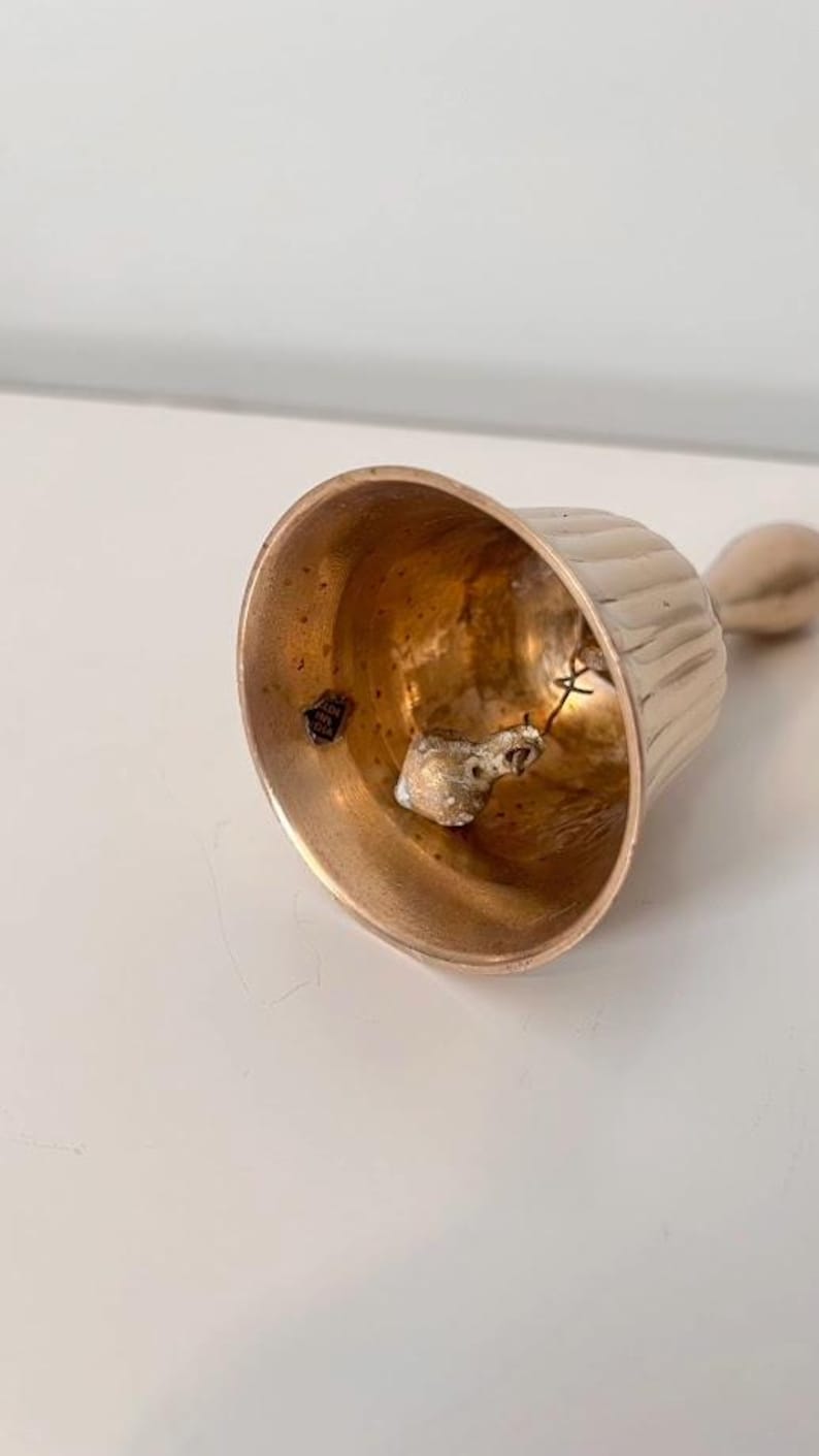 Pode incluir: Uma campainha de m&atilde;o de metal em tom dourado com um design exterior canelado. O interior da campainha apresenta um badalo suspenso por um fio e um pequeno autocolante que diz Made in India. A campainha tem uma pega lisa no topo.