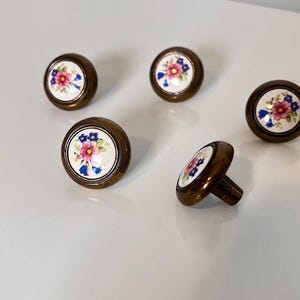 May include: Four round cabinet knobs featuring a white ceramic centre with a painted floral design of pink and blue flowers with green leaves. Each knob has a dark metallic bronze-toned rim and a matching metal base for mounting to furniture.