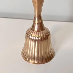 Pode incluir: Uma campainha de m&atilde;o em lat&atilde;o polido com um cabo liso e arredondado e um corpo alargado e canelado com cristas verticais. A superf&iacute;cie met&aacute;lica tem um tom dourado quente e reflexivo, exibindo um design cl&aacute;ssico com uma base circular larga.