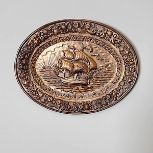 Pode incluir: Uma placa de parede oval em tom de bronze com um design em relevo de um veleiro na água com um sol nascente ao fundo. A borda é decorada com um padrão detalhado em relevo de várias frutas, nozes e folhagens.