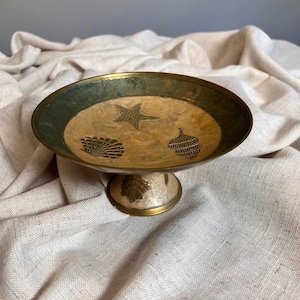 Pode incluir: Uma taça de latão sobre pedestal com uma base larga e rasa, uma borda metálica escura e um centro mais claro. A superfície interior é decorada com ilustrações gravadas de uma estrela-do-mar, uma vieira e uma concha em espiral.
