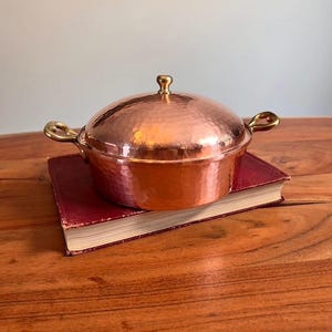 May include: A round, hammered copper casserole dish with a matching lid and two brass side handles. The lid features a small, rounded brass knob at the centre. The exterior surface of the pot and lid has a distinct, textured, hammered finish.