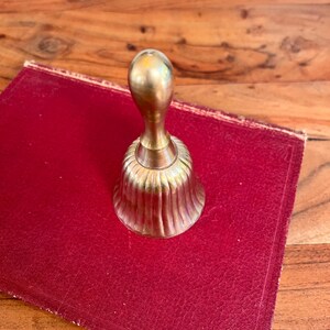 Pode incluir: Uma campainha de m&atilde;o em lat&atilde;o polido com um cabo liso e arredondado e um corpo canelado em forma de sino. A campainha apresenta um acabamento met&aacute;lico dourado com subtis estrias verticais ao longo da sec&ccedil;&atilde;o inferior.