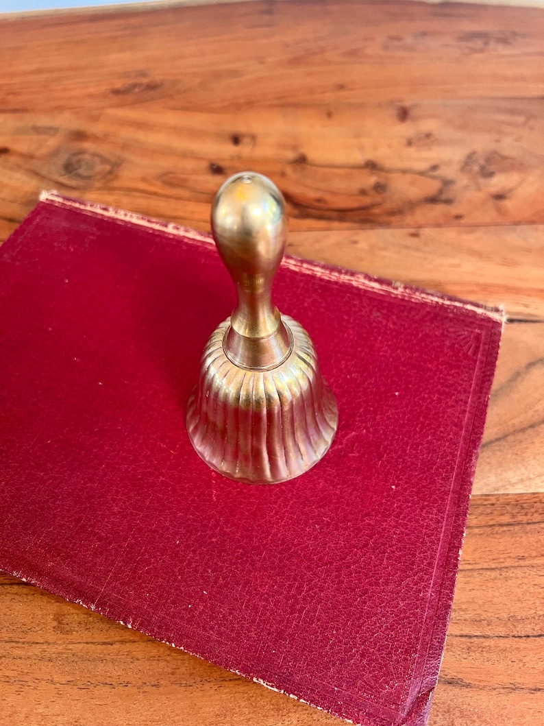 Pode incluir: Uma campainha de m&atilde;o em lat&atilde;o polido com um cabo liso e arredondado e um corpo canelado em forma de sino. A campainha apresenta um acabamento met&aacute;lico dourado com subtis estrias verticais ao longo da sec&ccedil;&atilde;o inferior.