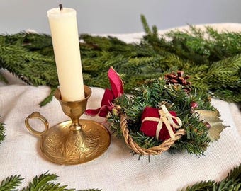 Peut inclure: Un bougeoir en laiton avec un motif floral en relief et une poignée latérale, contenant une bougie effilée couleur crème. À côté se trouve une petite couronne décorative en aiguilles de pin, un cadeau en velours rouge et un ruban rouge.