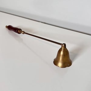 Pode incluir: Um apagador de velas de latão com um cabo de metal longo e fino e uma empunhadura de madeira castanho-escura na extremidade. O apagador possui uma cabeça em forma de sino com borda recortada, projetada para extinguir chamas de velas.