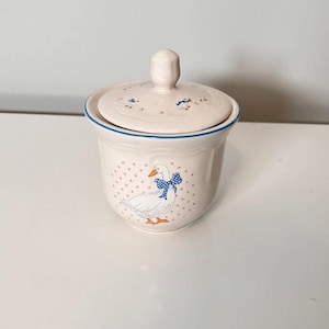 Può includere: Una zuccheriera in ceramica color crema con coperchio abbinato, caratterizzata da un'illustrazione di un'oca con un fiocco blu a pois, dettagli arancioni e un delicato motivo a pois rosa. Il bordo è rifinito con una sottile linea blu e il coperchio ha un pomello.