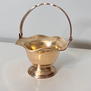 Pode incluir: Uma cesta de latão polido com borda recortada e base arredondada, apresentando uma alça de metal fina e arqueada fixada nas laterais com pequenos rebites. A superfície metálica reflete a luz, destacando a forma suave e curva do recipiente.