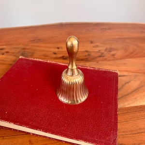 Pode incluir: Uma campainha de m&atilde;o em lat&atilde;o polido com um cabo liso e arredondado e um corpo canelado em forma de sino com estrias verticais. A campainha tem um acabamento met&aacute;lico dourado e permanece na vertical com uma base alargada e um pesco&ccedil;o estreito.
