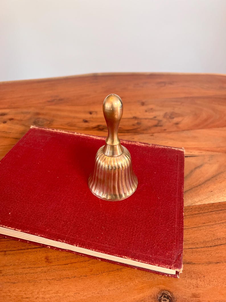 Pode incluir: Uma campainha de m&atilde;o em lat&atilde;o polido com um cabo liso e arredondado e um corpo canelado em forma de sino com estrias verticais. A campainha tem um acabamento met&aacute;lico dourado e permanece na vertical com uma base alargada e um pesco&ccedil;o estreito.